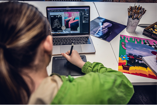 A designer using a stylus and drawing tablet while working on graphic design software on a MacBook laptop.